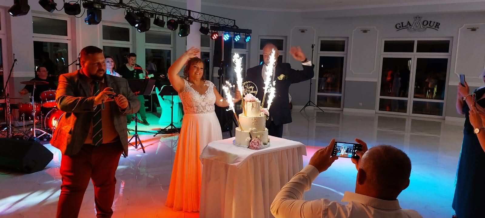 Sala Weselna Glamour Chroboty pod Żyrardowem - tort weselny z fontannami zimnych ogni, para młoda przy torcie podczas wieczornego przyjęcia, zespół muzyczny i logo Glamour na ścianie sali