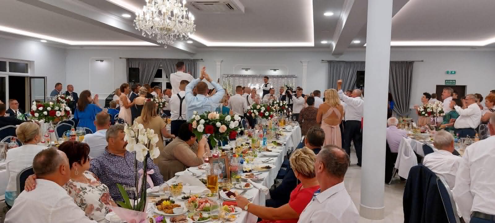 Sala Weselno-Bankietowa Glamour pod Żyrardowem - panorama pełnej sali podczas przyjęcia, długie stoły z dekoracją z białych i czerwonych róż, goście i kryształowy żyrandol