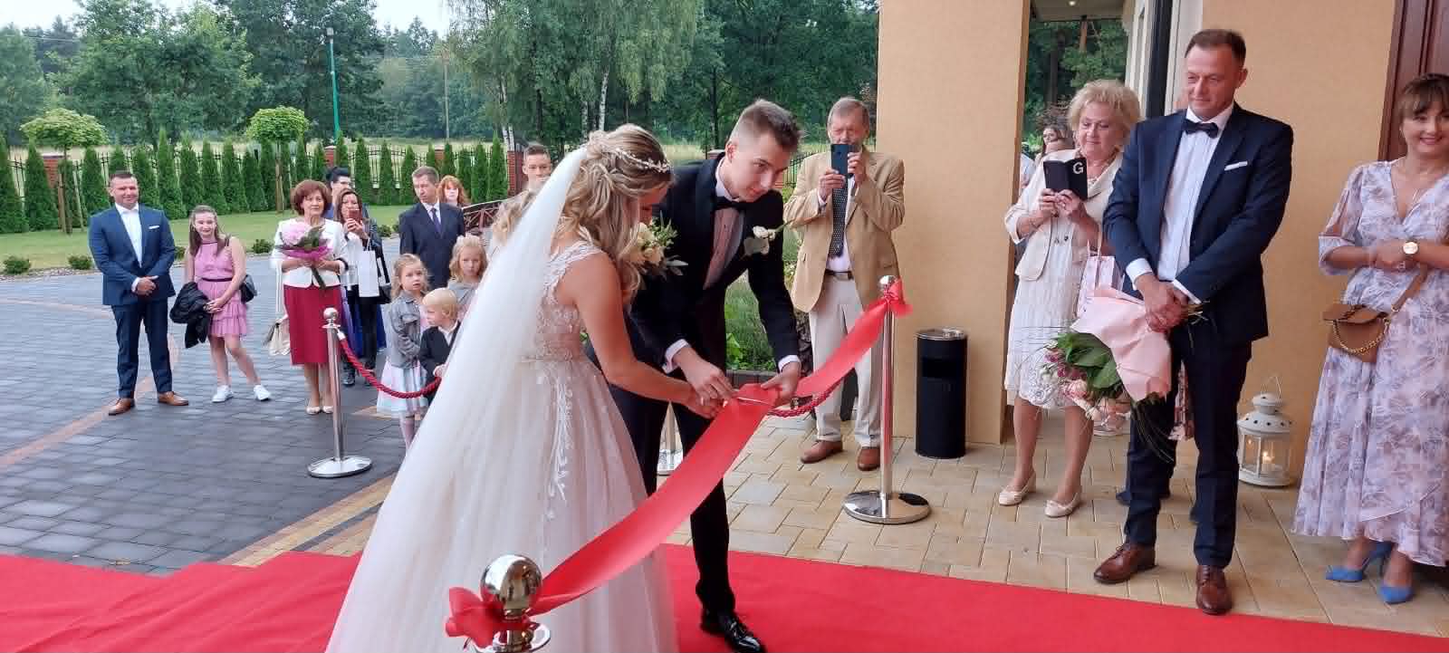 Sala Weselno-Bankietowa Glamour Chroboty - przecięcie wstęgi przez parę młodą przed wejściem do sali, czerwony dywan i goście witający nowożeńców